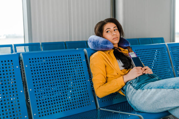 tired african american woman sitting with travel pillow and passport and air ticket  in departure lounge