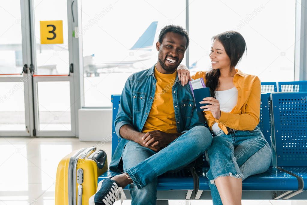 happy african american couple sitting in departure lounge with baggage and air tickets in airport