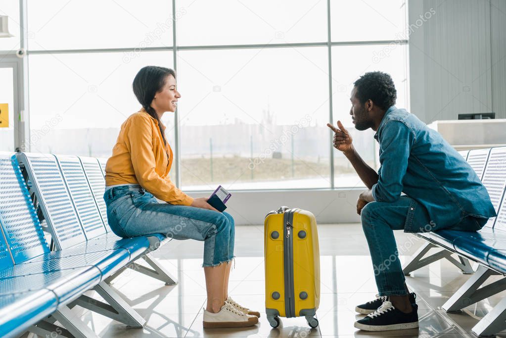 side view of african american couple sitting in departure lounge with suitcase and air tickets in airport