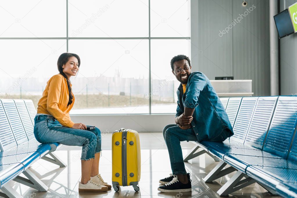 side view of happy african american couple sitting in departure lounge with suitcase in airport
