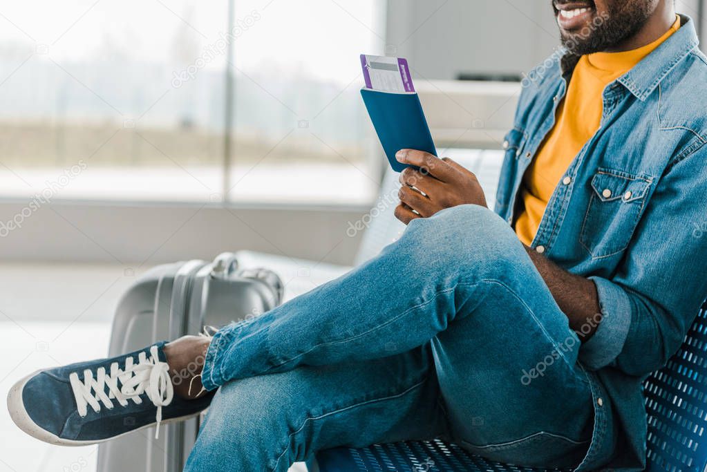 cropped view of smiling african american man sitting in airport with air ticket and passport