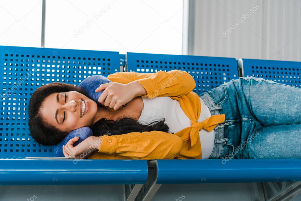 smiling african american woman lying on seats with travel pillow and closed eyes in departure lounge