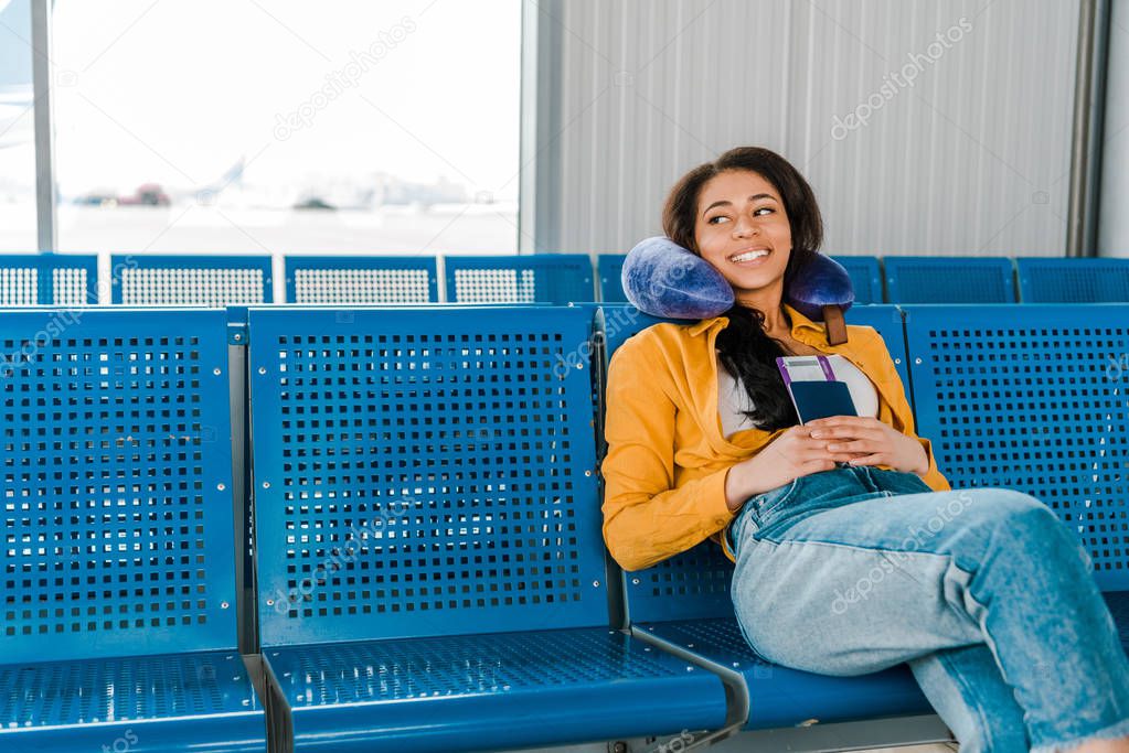 smiling african american woman sitting with travel pillow and passport and air ticket  in departure lounge