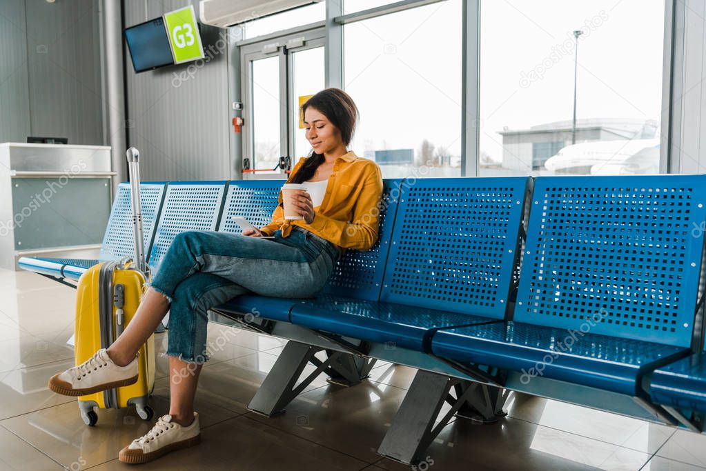 smiling african american woman sitting in departure lounge with suitcase, coffee to go and using smartphone