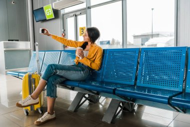 gülümseyen African Amerikan kadın bavul ile ayrılış salonunda oturan, kahve gitmek ve akıllı telefonda selfie almak
