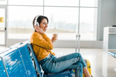 kulaklık müzik dinlerken mutlu Afrikalı Amerikan kadın gitmek ve bavul kahve ile ayrılış salonunda oturan 