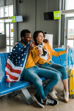 havaalanında ayrılış salonunda Amerikan bayrağı ve Bagaj ile oturan mutlu Afrikalı Amerikan çift 