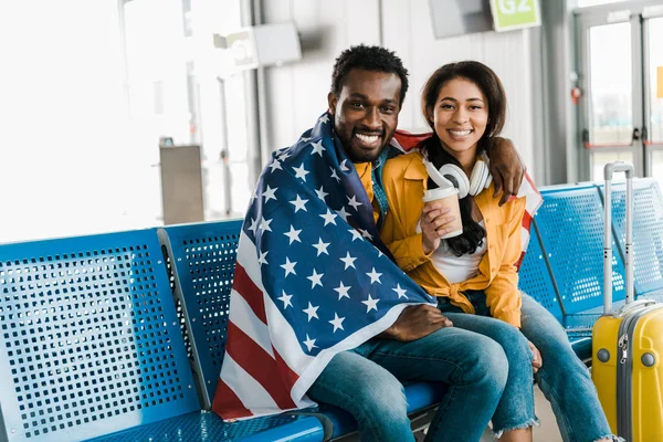 smiling african american couple sitting with american flag in departure lounge in airport