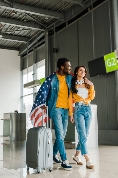 happy african american couple with american flag and suitcase walking  in departure lounge in airport 