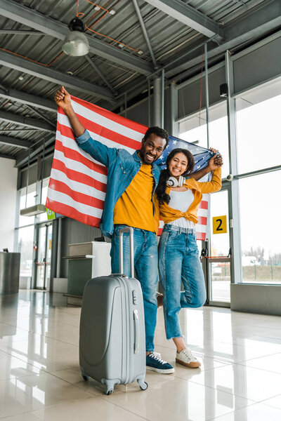 smiling african american couple with american flag and luggage walking  in departure lounge in airport 