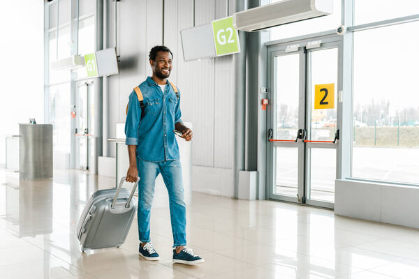 счастливый красивый африканский американец гуляющий с чемоданом в аэропорту
