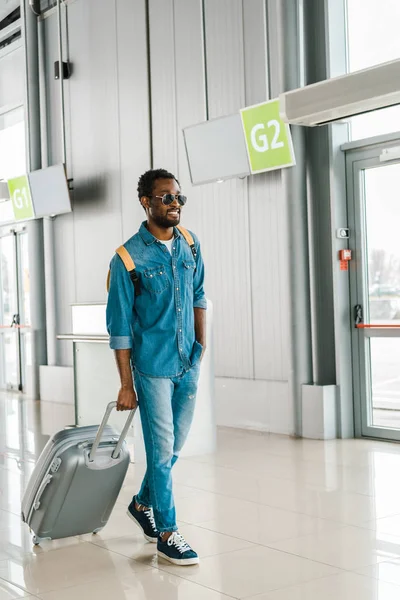 サングラスでハンサムなアフリカ系アメリカ人の男性を幸せにする空港でスーツケースを歩いて — ストック写真