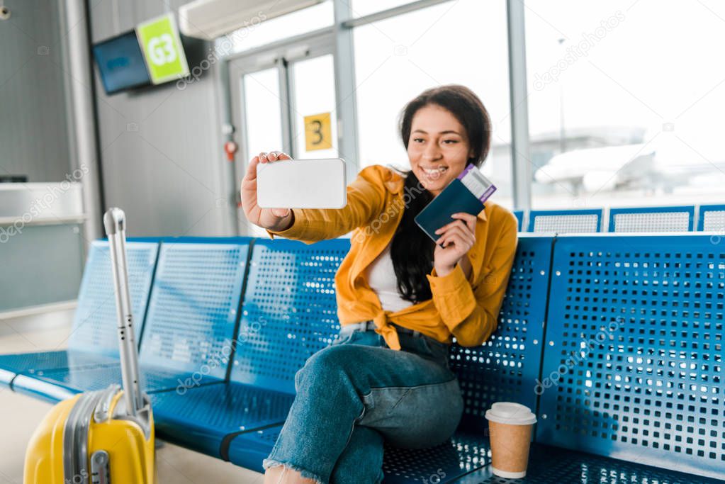 happy african american woman sitting in departure lounge with suitcase, coffee to go and taking selfie with passport and air ticket on smartphone