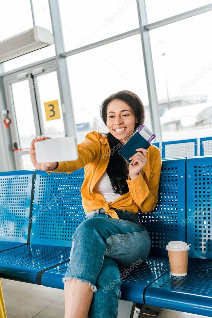 happy african american woman sitting in departure lounge and taking selfie with passport and air ticket on smartphone