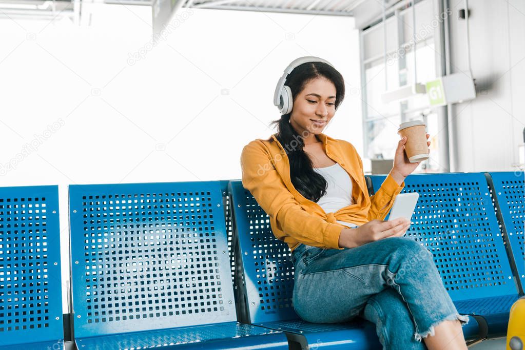 happy african american woman sitting in departure lounge with coffee to go while listening music in headphones and using smartphone
