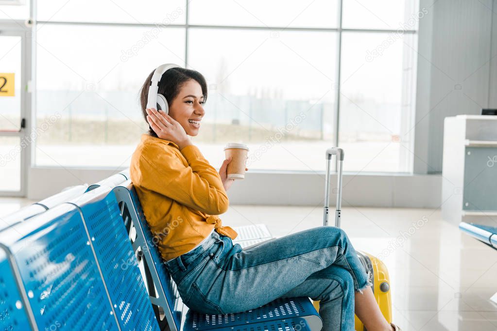 happy african american woman sitting in departure lounge with coffee to go and suitcase while listening music in headphones 