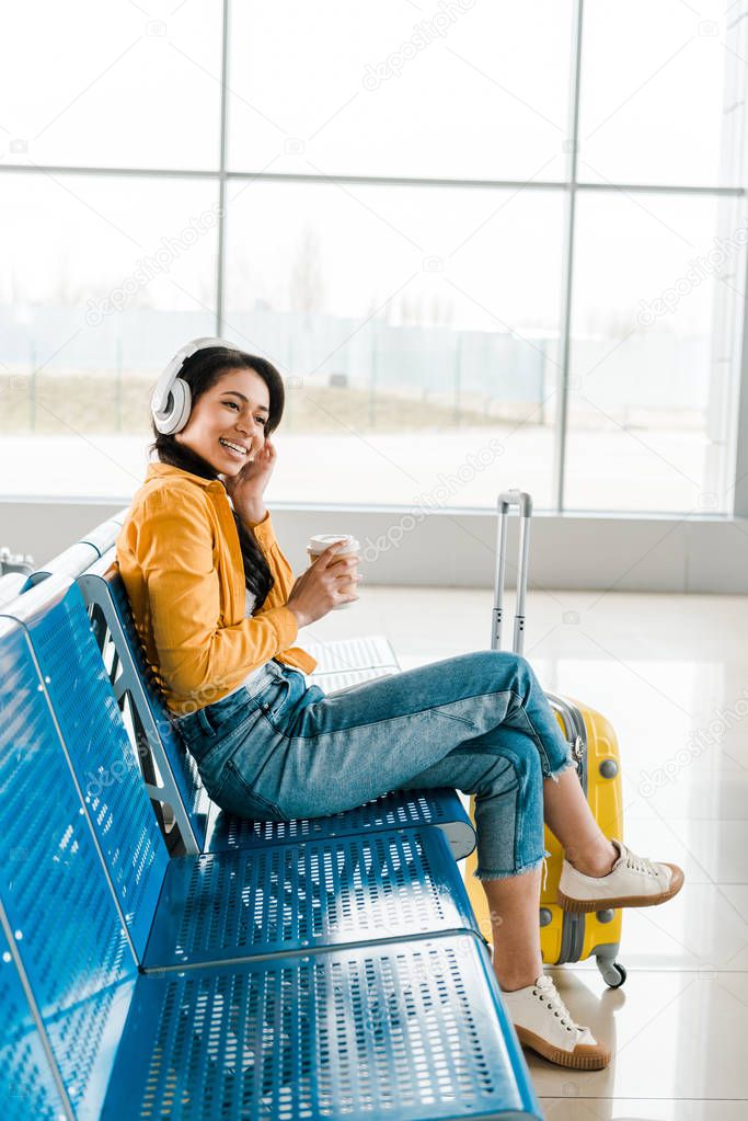 smiling african american woman sitting in departure lounge with coffee to go and suitcase while listening music in headphones 
