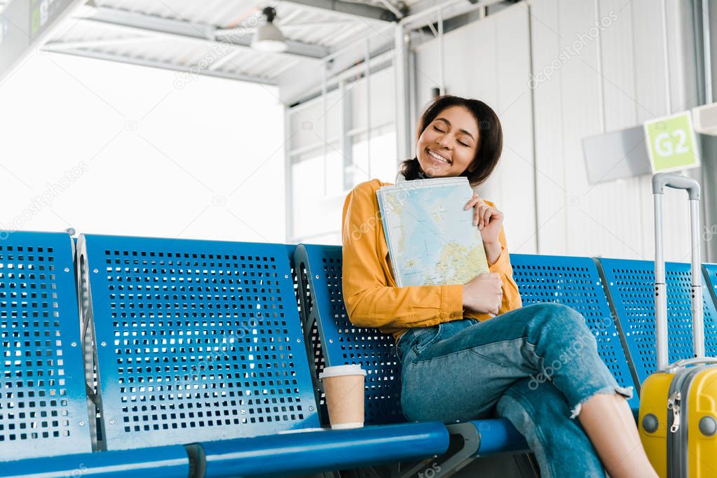 smiling african american woman sitting with coffee to go and map in departure lounge in airport