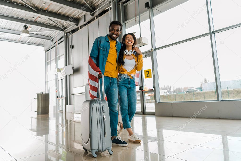 happy african american couple with american flag and luggage walking  in departure lounge in airport 