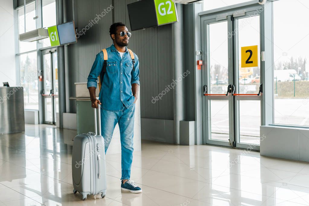 handsome african american man standing with hand in pocket and suitcase in airport