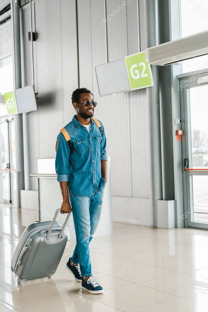 happy handsome african american man in sunglasses walking with suitcase in airport