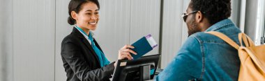 panoramic shot of smiling african american airport worker giving passport and air ticket to tourist with backpack clipart
