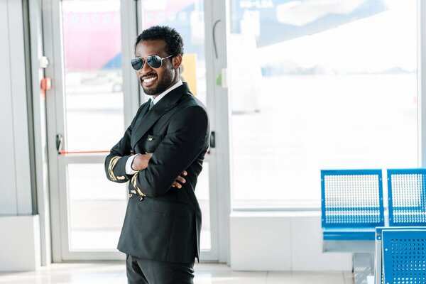smiling african american pilot standing with arms crossed in departure lounge in airport
