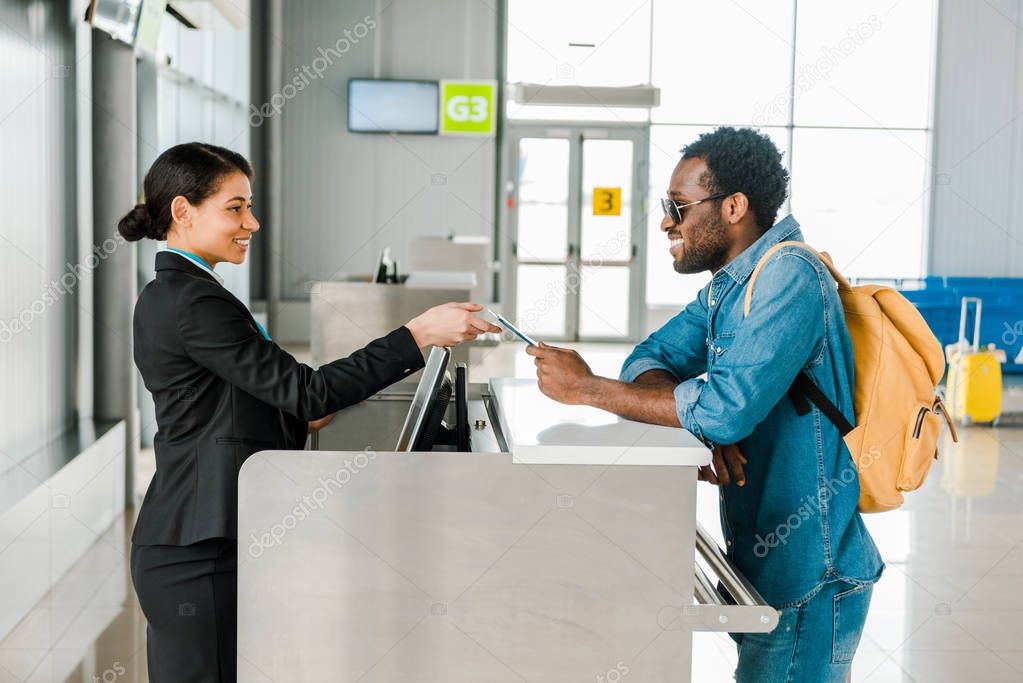 smiling african american airport worker taking passport and air ticket from tourist with backpack