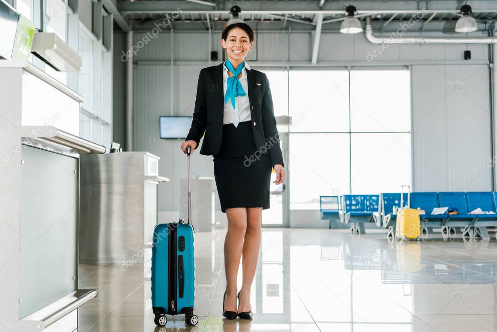 smiling african american stewardess standing with suitcase in airport