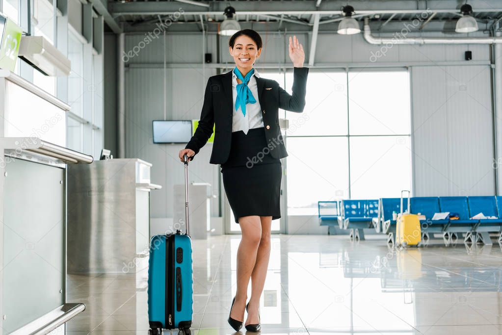 smiling african american stewardess with travel bag waving hand in airport