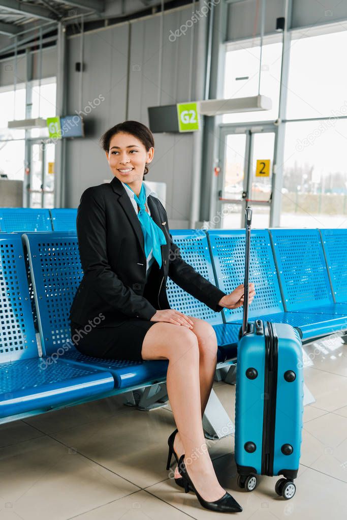 smiling african american stewardess sitting with suitcase in departure lounge