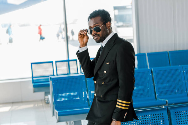 stylish african american pilot in sunglasses near seats in departure lounge in airport