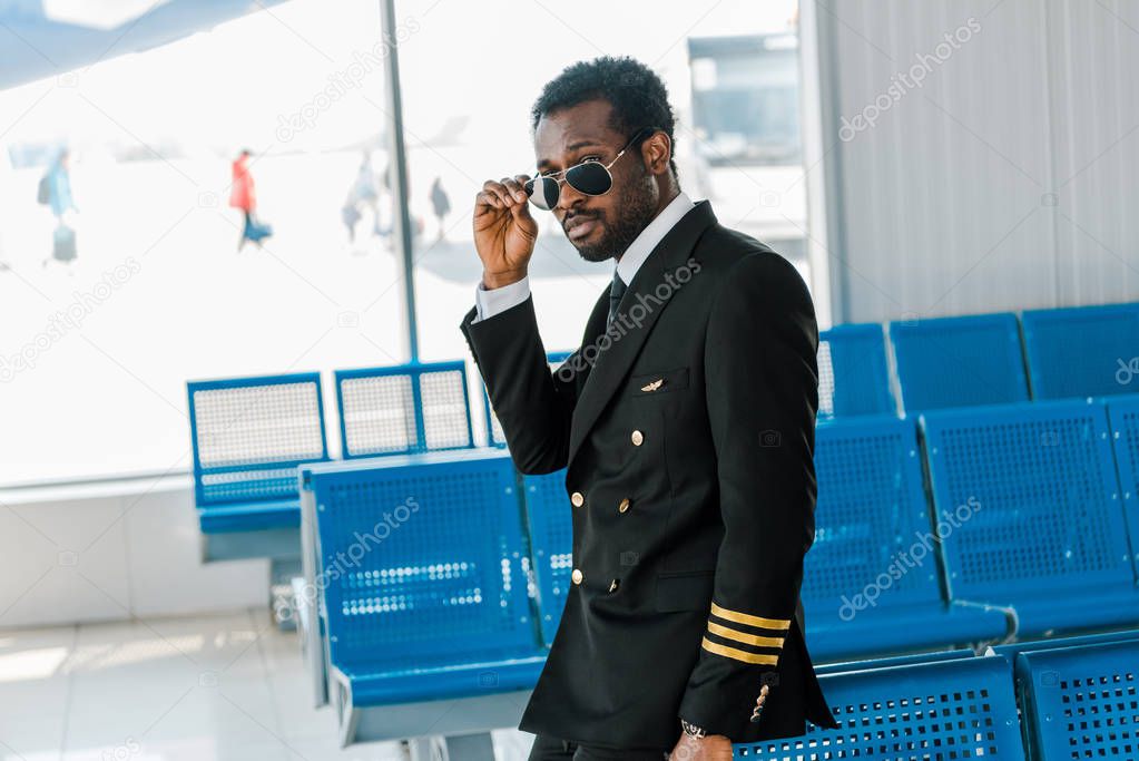 stylish african american pilot in sunglasses near seats in departure lounge in airport