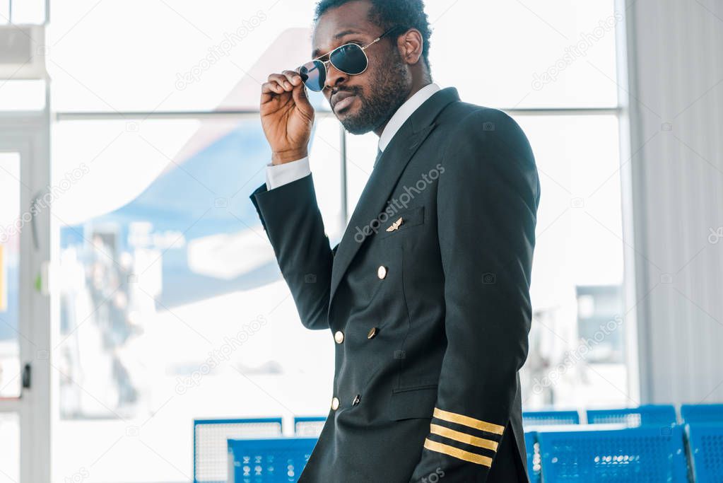 stylish confident african american pilot in sunglasses in departure lounge in airport