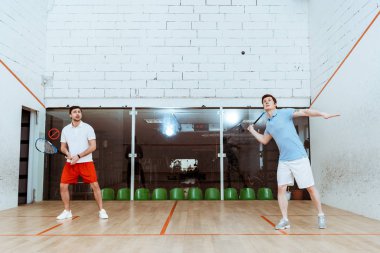 Dört duvarlı kortta raketlerle squash oynayan iki sporcunun tam boy görünümü