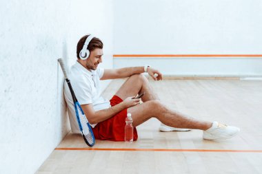 Smiling squash player listening music in headphones and using smartphone clipart