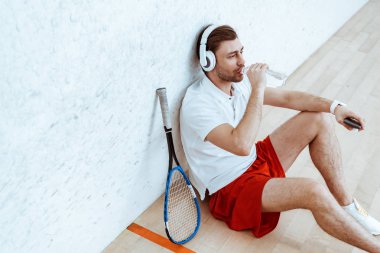 High angle view of squash player listening music in headphones and drinking water clipart