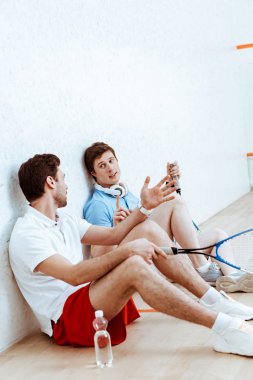 Dört duvarlı sahada yerde otururken iki squash oyuncusu konuşuyor