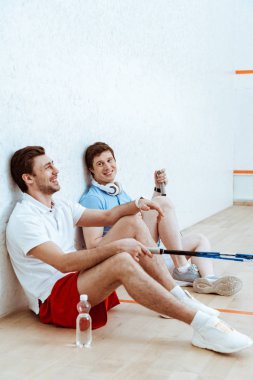 Dört duvarlı sahada yerde oturan iki gülümseyen squash oyuncusu