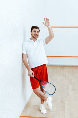 Raket tutan ve el sallayarak kırmızı şort squash oyuncu tam uzunlukta görünümü