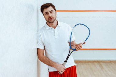 Sakallı squash oyuncu raket tutan ve kamera bakıyor