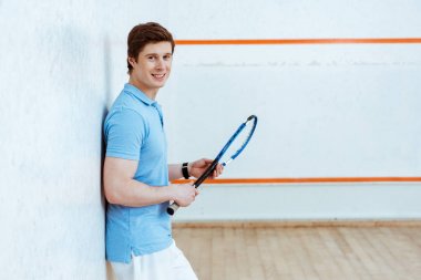 Mavi polo gömlek raket tutan ve kameraya bakarak gülümseyen squash oyuncu