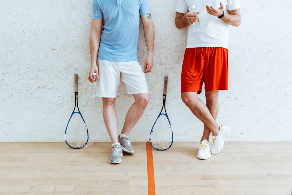 Cropped view of two squash players in shorts standing with crossed legs in four-walled court
