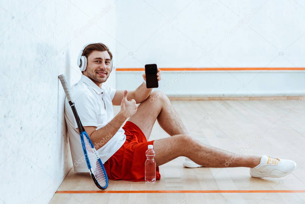 Squash player in headphones showing thumb up and holding smartphone with blank screen