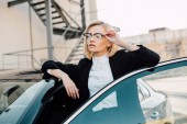 sebevědomá blondýnka v brýlích stojících poblíž černého auta a dotýkala se brýlí  