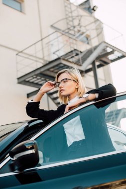 siyah otomobil ve dokunaklı gözlük yakınında duran gözlük emin sarışın genç kadın düşük açı görünümü  