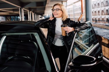 gözlük neşeli kız akıllı telefon konuşurken ve araba yakınında tek kullanımlık fincan tutarak 