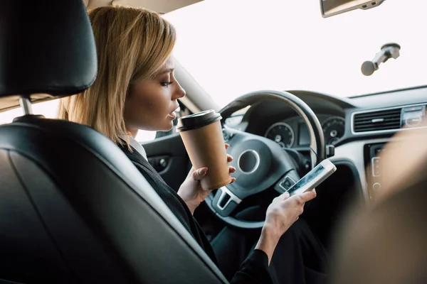 車の中でスマートフォンを見ながら紙コップを持つブロンドの若い女性 — ストック写真