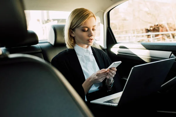 有吸引力的金发年轻女子看着智能手机 而坐在笔记本电脑在汽车 — 图库照片