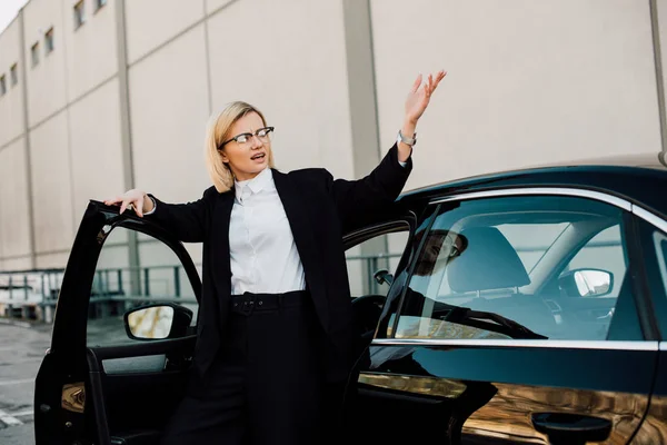 offended blonde young woman in glasses standing near black automobile and gesturing in parking